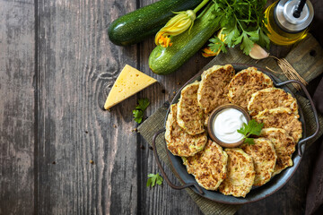Healthy summer food, zucchini fritters. Vegetarian zucchini pancakes with cheese, served with sour cream on a rustic wooden table. Top view flat lay background.