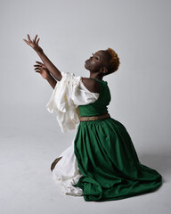 Full length portrait of pretty African woman wearing long green medieval fantasy gown, kneeling pose on a light grey studio background.