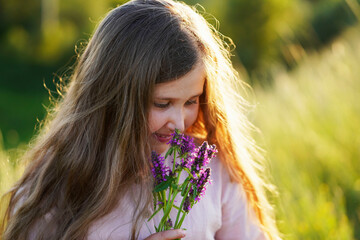 Happy cute funny smiling caucasian girl walking on the meadow and holding a bouquet of wildflowers.  Summer sunset.  Happy Childhood concept. Nature background. Copy space