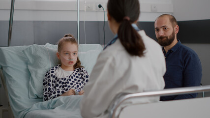 Obraz premium Pediatrician doctor discussing sickness treatment with sick patient and father during disease medicine examination in hospital ward. Hospitalized child lying in bed recovering after medical surgery