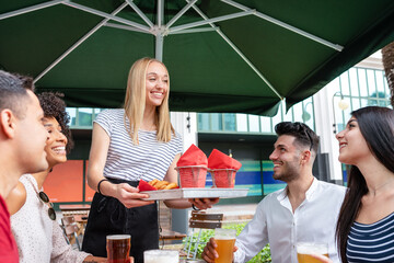 young blonde waitress serving food and french fries, two couple at the table waiting their order in a pub, people eating at restaurant and drinking beer, youth lifestyle
