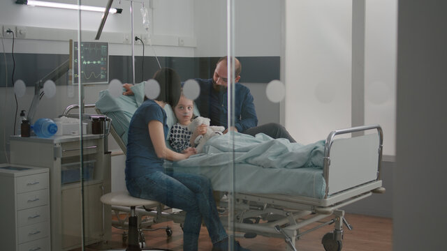 Worried parents sitting with sick girl daughter waiting for sickness expertise during medical examination in hospital ward. Child wearing oxygen nasal tube holding teedy bear while resting in bed