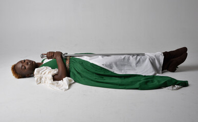 Full length portrait of pretty African woman wearing long green medieval fantasy gown, sitting on a light grey studio background. © faestock