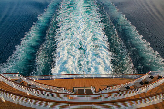 Looking down at the aft or back decks of a cruise ship and the wake or waves in the sea as it sails across the ocean. Open decks with tables and chairs and railings to view and relaz.