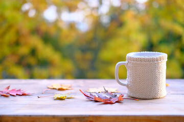Naklejka na ściany i meble beverage, coffee in a mug, leaves, foliage on the wooden table in the garden, beautiful blurred natural landscape in the background, good weather concept, outdoor tea party, cozy autumn mood