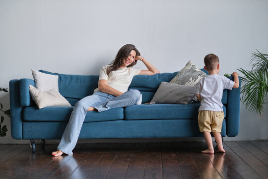 Tired Young Mother Or Nanny Sits On The Couch And Watches Her Son Playing.