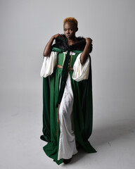 Full length portrait of pretty African woman wearing long green medieval fantasy gown and velvet cloak, sitting on a light grey studio background.