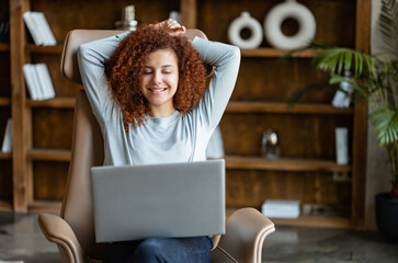 Serene carefree attractive red-haired curly woman using laptop sitting on the cozy chair, holding arms behind head, resting at home, watching movie, spends leisure in networks