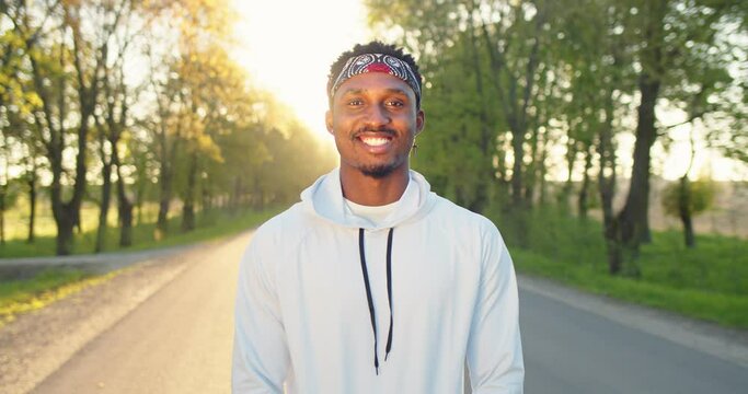 Portrait of happy young African American smiling handsome male athlete looking at camera in good mood while standing on sunny street outdoor on sunset. Positive sportsman in park in town