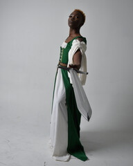 Full length portrait of pretty African woman wearing long green medieval fantasy gown holding  sword, standing action pose on a light grey studio background.