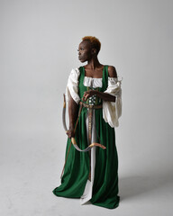 Full length portrait of pretty African woman wearing long green medieval fantasy gown holding bow and arrow, standing action pose on a light grey studio background.