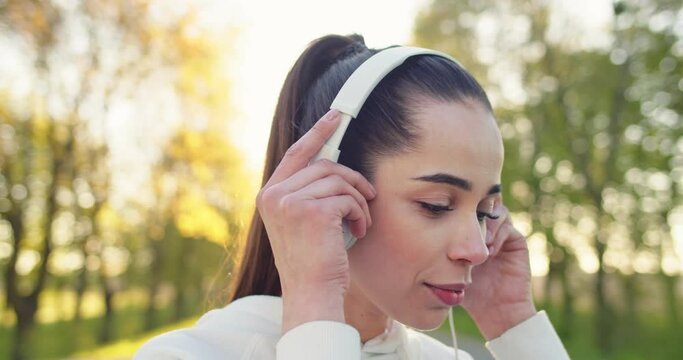 Close up of cheerful smiling young beautiful Caucasian sportswoman standing outdoors putting headphones on and listening to music before workout. Leisure, recreation, urban life concept