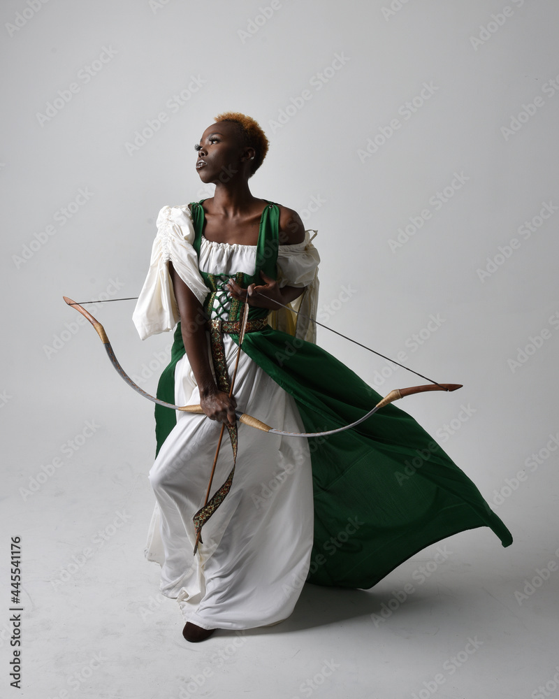 Poster Full length portrait of pretty African woman wearing long green medieval fantasy gown holding bow and arrow, standing action pose on a light grey studio background.