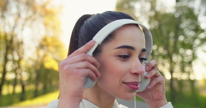 Close up shot of cheerful young Caucasian pretty sportswoman standing outdoors putting headphones on and looking at camera smiling while listening to music. Leisure concept, city urban
