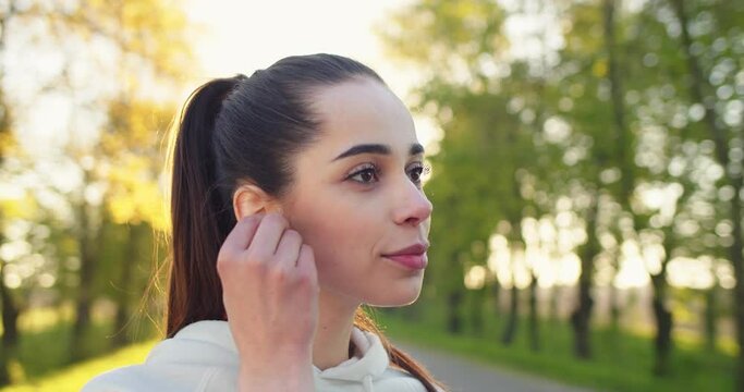 Close up of happy positive young Caucasian beautiful sportswoman standing outdoors alone putting wireless earphones on and listening to music in good mood. Leisure concept, park, recreation