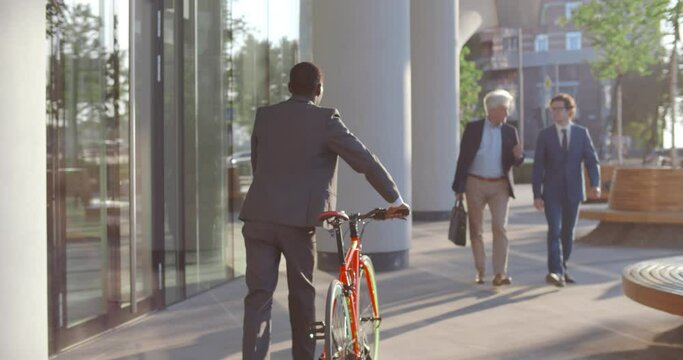Follow shot of african businessman with bicycle walking near business center