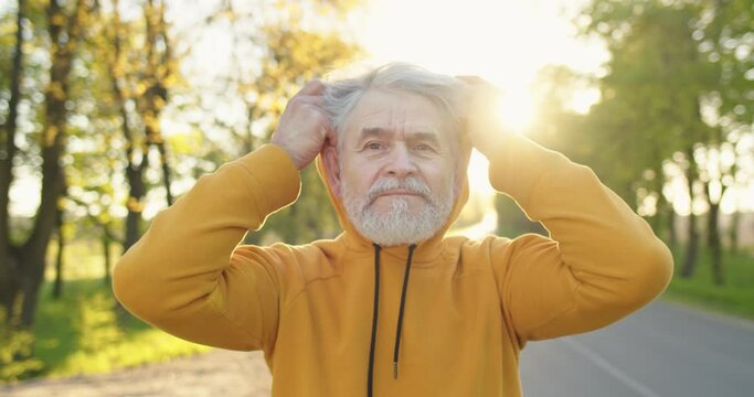 Close up portrait of happy Caucasian handsome old man in good mood putting on hood and looking at camera standing on street outdoors. Senior male on a walk in park, outdoor, recreation concept