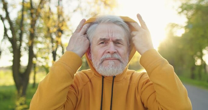 Close up portrait of Caucasian handsome old joyful man in good mood putting on hood and looking at camera standing on street alone outdoors in sun lights Senior male on a walk in park recreation sport