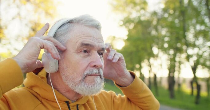 Close up of happy senior Caucasian handsome grey-haired bearded man standing outdoors alone putting headphones on and listening to music. Leisure concept, park, recreation, walk, sport