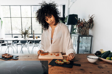 Young multiracial woman with curly hair preparing avocado toast for breakfast in big bright kitchen