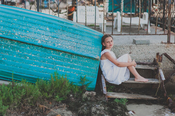A charming teenager girl with long hair in a white dress sits leaning against a turquoise wooden boat. Mood spleen