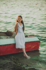 Pin-up girl teenager with long hair in a white dress stands leaning against a fishing boat in the...