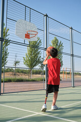 Back view of a basketball kid player with a leg prosthesis throwing the ball to the basket....