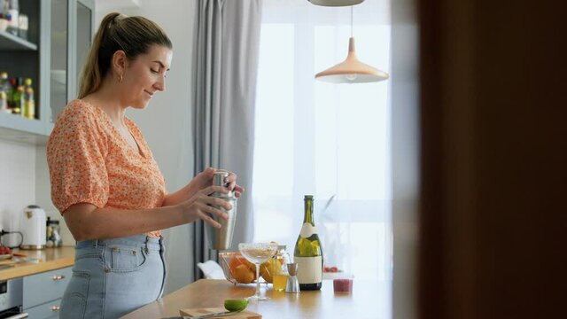 drinks and people concept - young woman with shaker pouring cocktail to glass at home kitchen