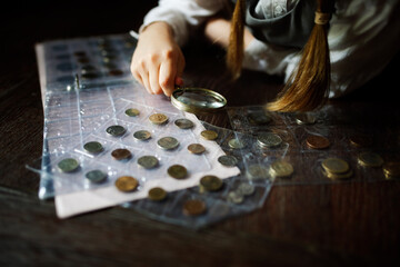Cute cute european girl child in glasses with magnifying glass looks at coins, concept children and financial literacy, children and money, coin collecting and numismatics, dark style - obrazy, fototapety, plakaty