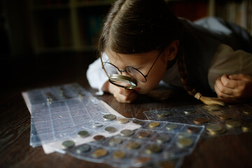 Cute cute european girl child in glasses with magnifying glass looks at coins, concept children and financial literacy, children and money, coin collecting and numismatics, dark style - obrazy, fototapety, plakaty