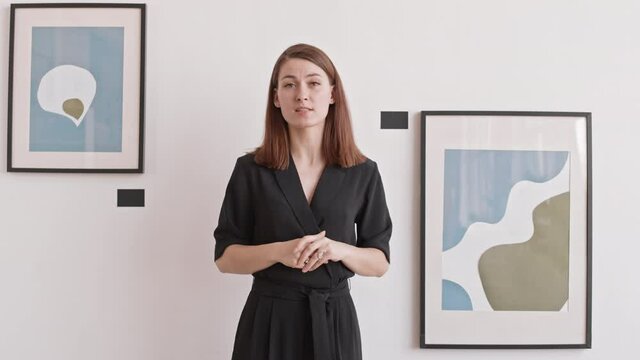 Medium of young Caucasian woman wearing total black outfit talking on camera, hosting online presentation of art exhibition in gallery