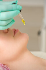 Close up of beautician holding pipette and applying oil serum drop on the face of a young woman in spa salon