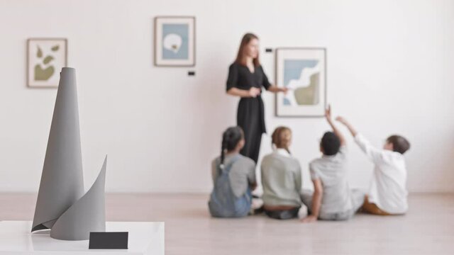 Racking focus from art object on pedestal to children sitting on floor of museum, listening to Caucasian woman talking about paintings on wall