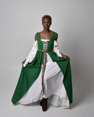 Fototapeta premium Full length portrait of pretty African woman wearing long green medieval fantasy gown, standing and dancing pose on a light grey studio background.