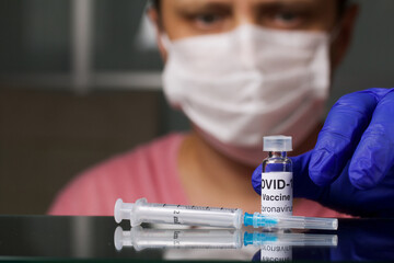 Vaccination against coronavirus. A man in a medical mask and gloves takes an ampoule with a vaccine. On the shelf is a syringe for injection. Close-up shot.
