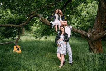 Fototapeta premium Two little sisters sitting on branch of old tree while father gently embracing pregnant mother. Caucasian family waiting for new addition.