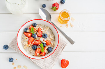 Bowl of oatmeal porridge with blueberries, strawberries, almond petals and honey on a white wooden background. Healthy food for breakfast, top view, copy space.