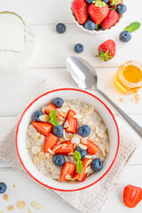 Bowl of oatmeal porridge with blueberries, strawberries, almond petals and honey on a white wooden background. Healthy food for breakfast, top view, copy space.