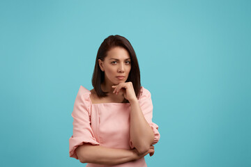 Obraz premium Pensive caucasian woman with dark hair wearing pink dress is thinking of something important, needs to make decision, looks at the camera, touches chin with her hand, isolated on blue background.