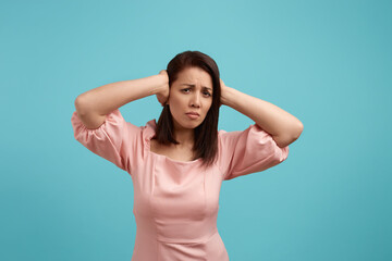 Fototapeta premium Attractive caucasian woman with dark hair wearing pink dress covers ears with hands not wanting to hear loud sound, wants calm and silence, protects herself from noise, isolated on blue background.