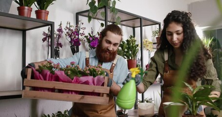 Diverse flower shop employees replanting various plants together. Multiethnic couple working at greenhouse - small business people, teamwork 4k footage - Powered by Adobe