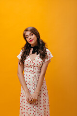 Curly woman in white dress smiling on orange background. Young shy girl with wavy hair in cherry print clothes looking into camera..
