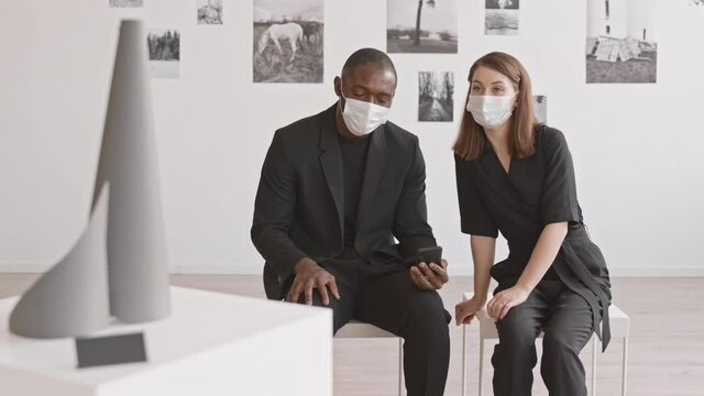 Medium long of young African man and Caucasian woman wearing black clothing and medical masks, sitting on stools in modern art gallery, talking and using smartphone