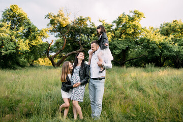 Fototapeta premium Young parents with two kids posing among high grass at green garden. Pregnant woman embracing older daughter while man holding on shoulders youngest one.