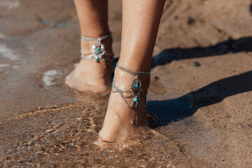 Close up of the feet in boho jewelry of a woman walking on the beach barefoot. Sunny summer day....