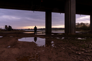 A bleak, moody, winter edit of a figure standing next to a lake, looking out under a motorway bridge at sunset