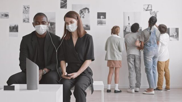 Medium long of young African man and Caucasian woman wearing black clothes and medical masks, sitting in modern art gallery, listening to audio guide in earphones, children standing on background