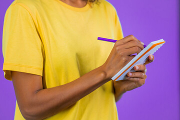 YHands of young woman making notes in planner, african lady holding pen. She writes future plans and to-do list in notebook for week or month. Keeping personal diary on purple studio background.