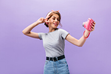 Dark-haired woman shows peace sign and holds camera. Cool cute girl in grey t-shirt and denim skirt with black belt posing..