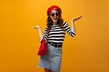 Fototapeta na wymiar Portrait of woman in red beret and striped shirt shrugging on orange background. Modern girl in sunglasses with modern handbag has fun..